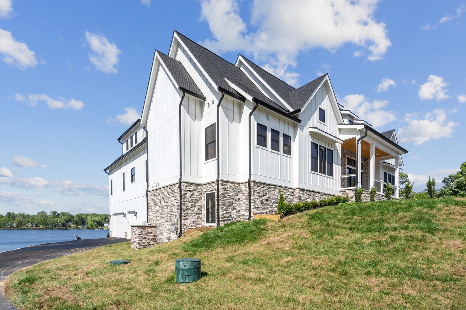 A large, modern white house with dark roofing stands on a grassy slope next to a lake under a partly cloudy sky.