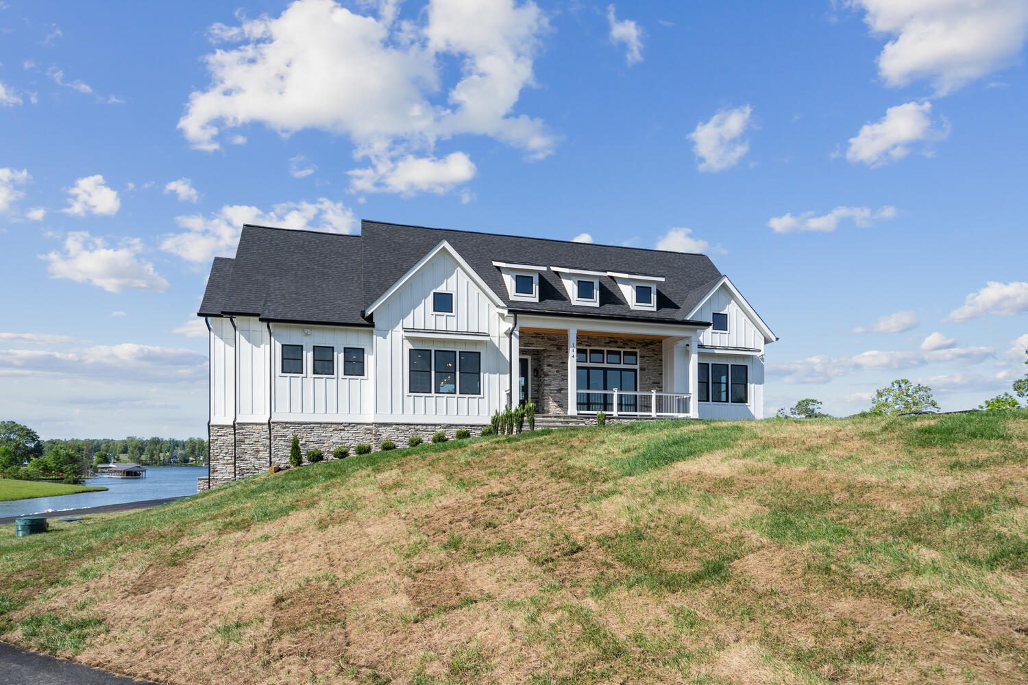 A modern farmhouse with a gray roof and white siding sits on a grassy hill near a lake, under a blue sky with clouds.