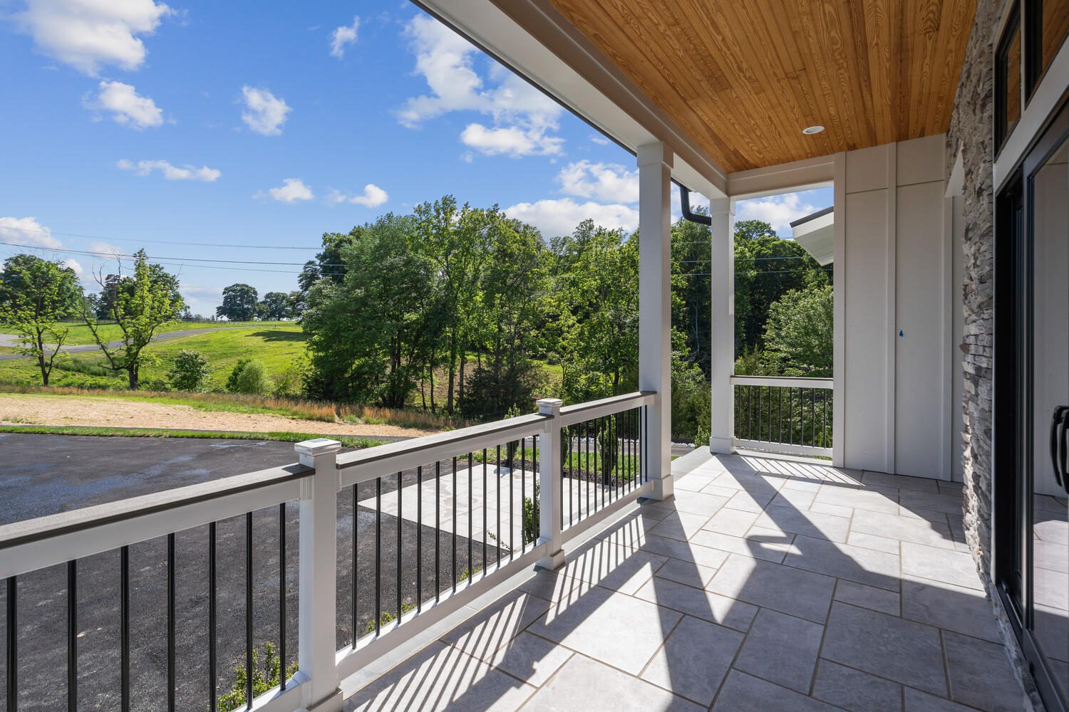 Covered porch with white railing overlooks a scenic view of a grassy field and trees under a blue sky with clouds. The floor is tiled, and the ceiling is wood-paneled.