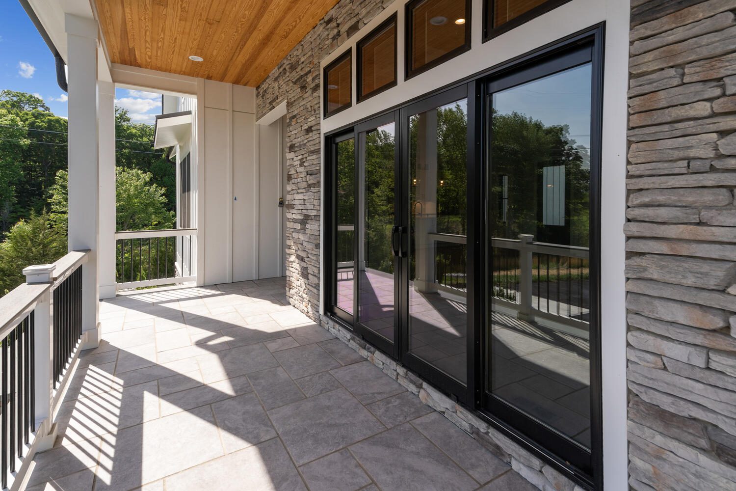 A patio with a stone wall, large glass sliding doors, and a tiled floor, overlooking a wooded area.