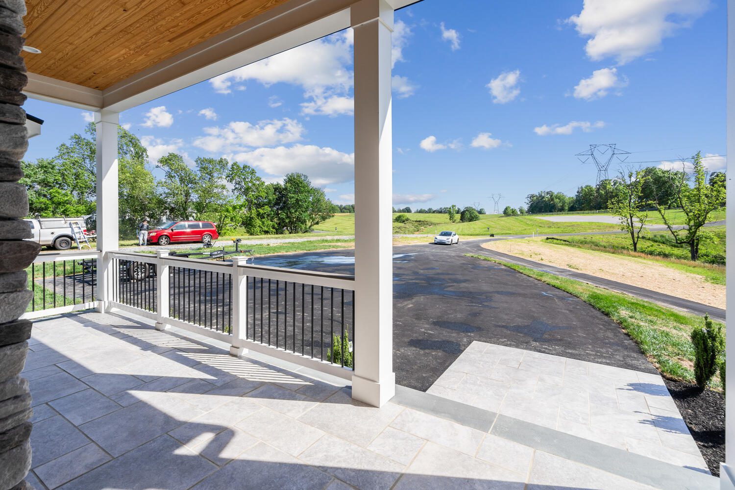 View from a porch with white railings, showing a curving driveway, grassy fields, a red truck, and a distant white car under a partly cloudy sky.