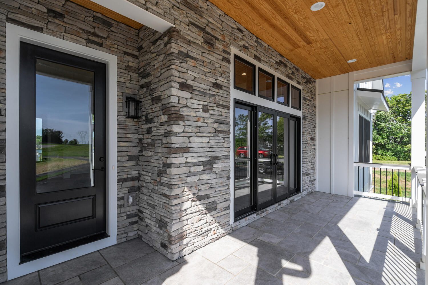 Stone-clad porch with large glass door and windows, overlooking a grassy landscape. The ceiling features wood paneling, and the floor is tiled in a light gray shade.