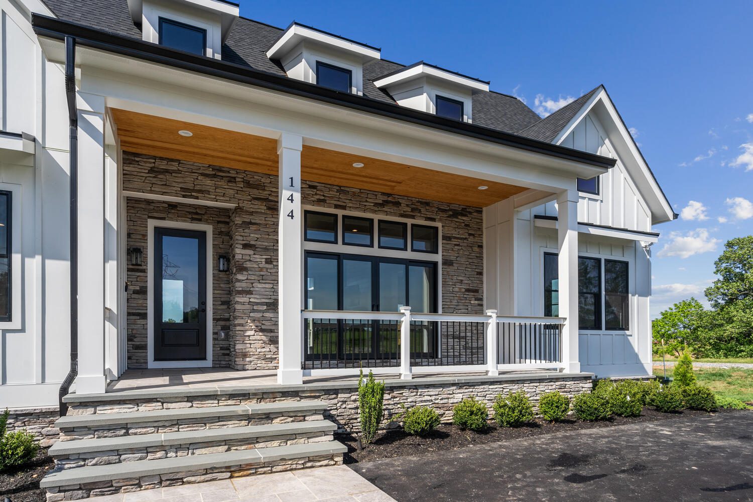 Modern house exterior with stone facade, large windows, a wooden porch ceiling, black-framed door, and small shrubs along the foundation. Steps lead up to the covered entryway.