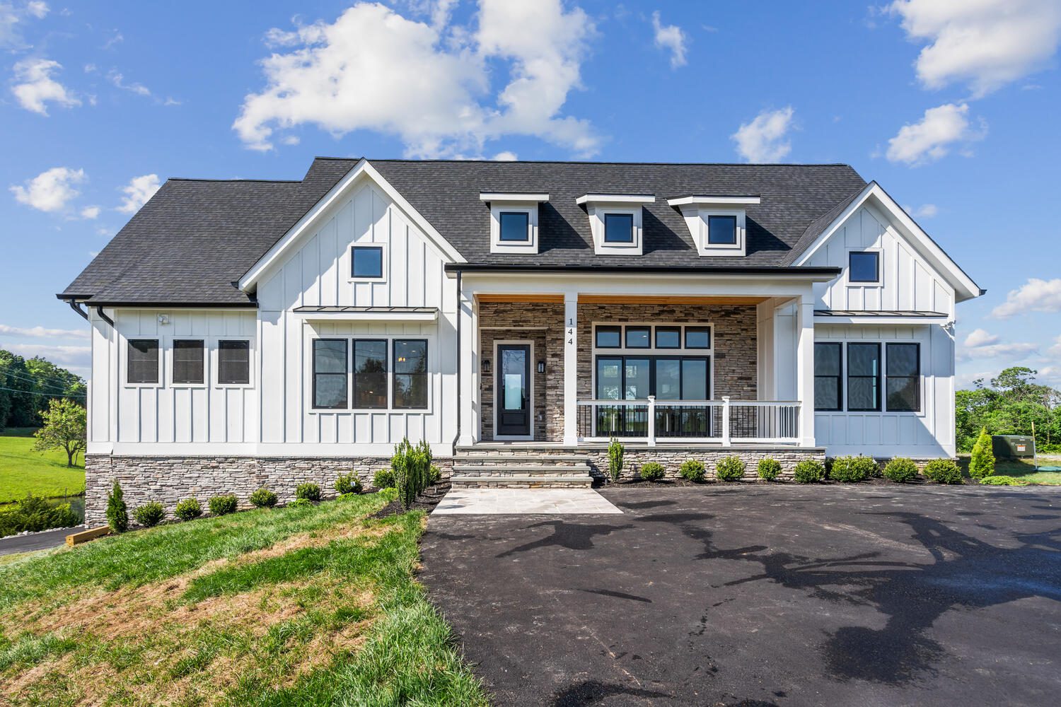 A large, modern house with white siding, stone accents, and a black roof. It features a front porch, multiple windows, and a neatly landscaped front yard under a blue sky with clouds.