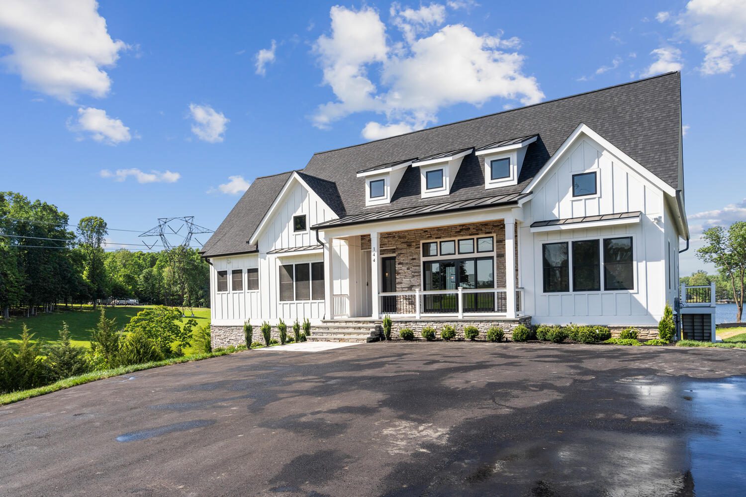 A modern two-story house with white siding and dark roof, featuring a front porch, large windows, and a paved driveway, surrounded by trees under a clear blue sky.