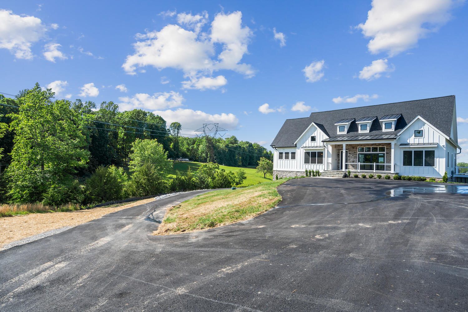 Large modern house with black roof and white facade, situated on a hill with surrounding greenery, under a partly cloudy sky. Freshly paved driveway in the foreground.