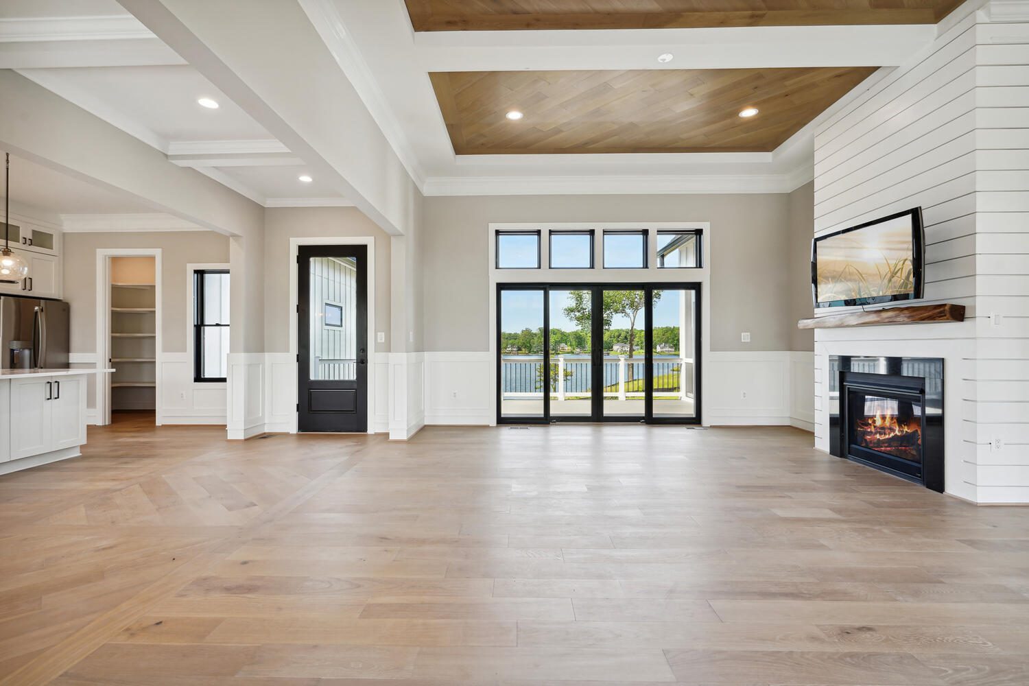 Spacious living room with hardwood floors, a fireplace, and large windows overlooking a balcony with a view. Ceiling features wood accents and recessed lighting. Kitchen visible in the background.