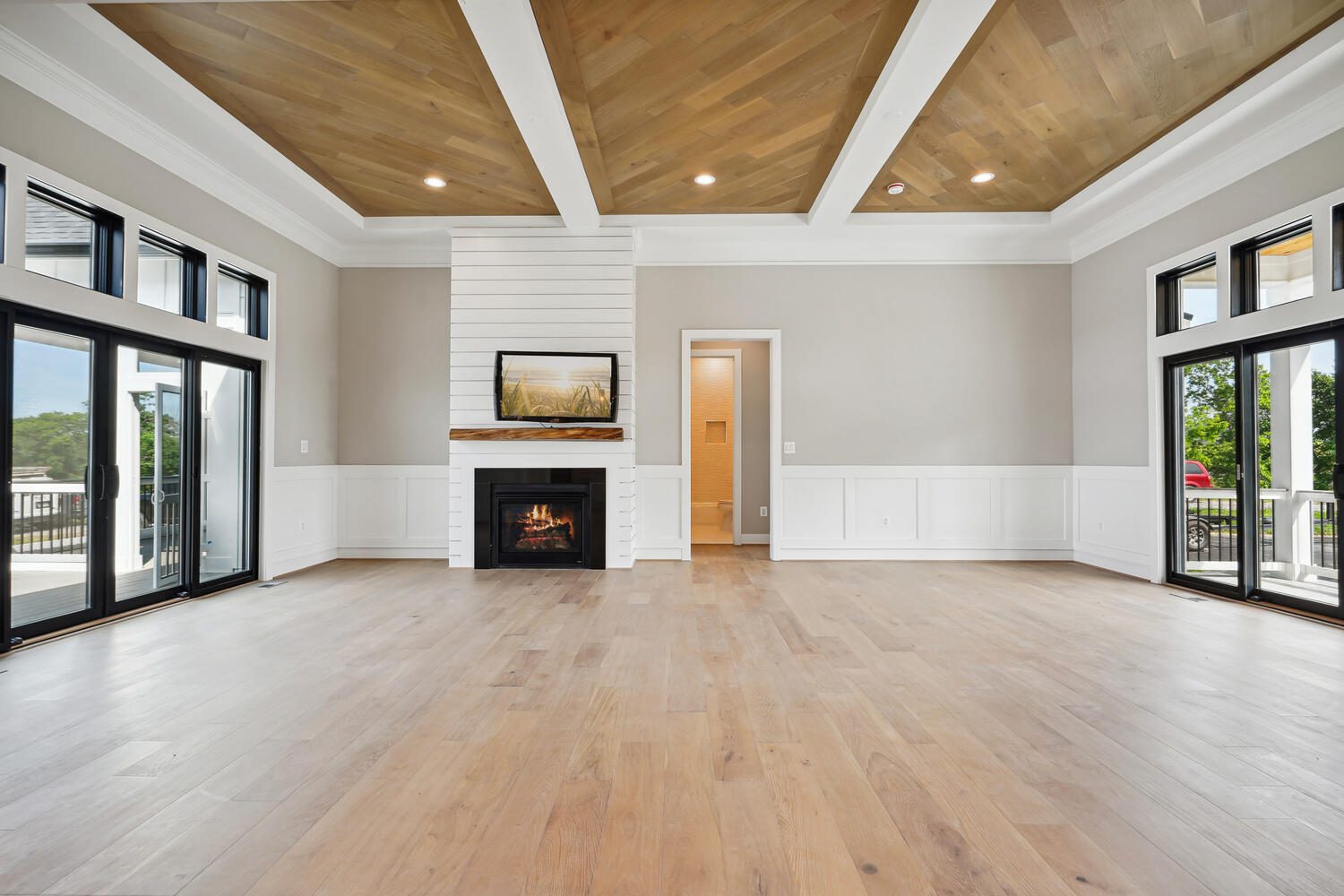 Spacious living room with wooden ceiling and floor, large windows, a fireplace, artwork above, and an open doorway leading to another area.