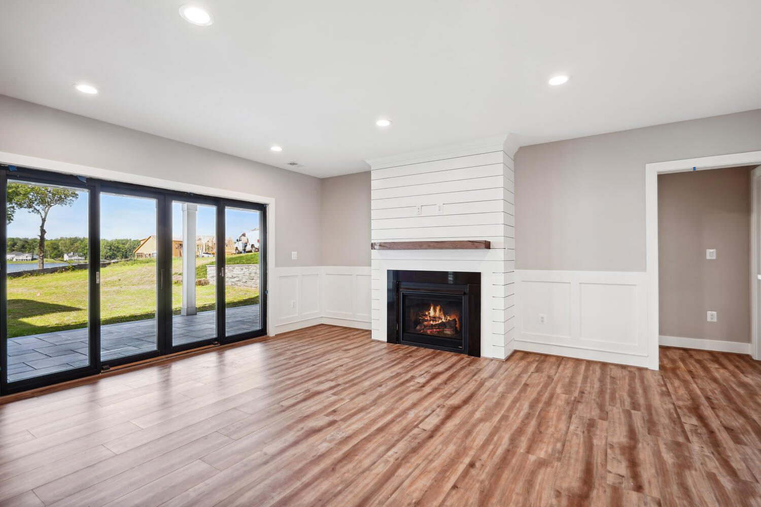 A spacious living room with wooden flooring, a fireplace, large sliding glass doors opening to a lawn, and white shiplap panels on the walls.