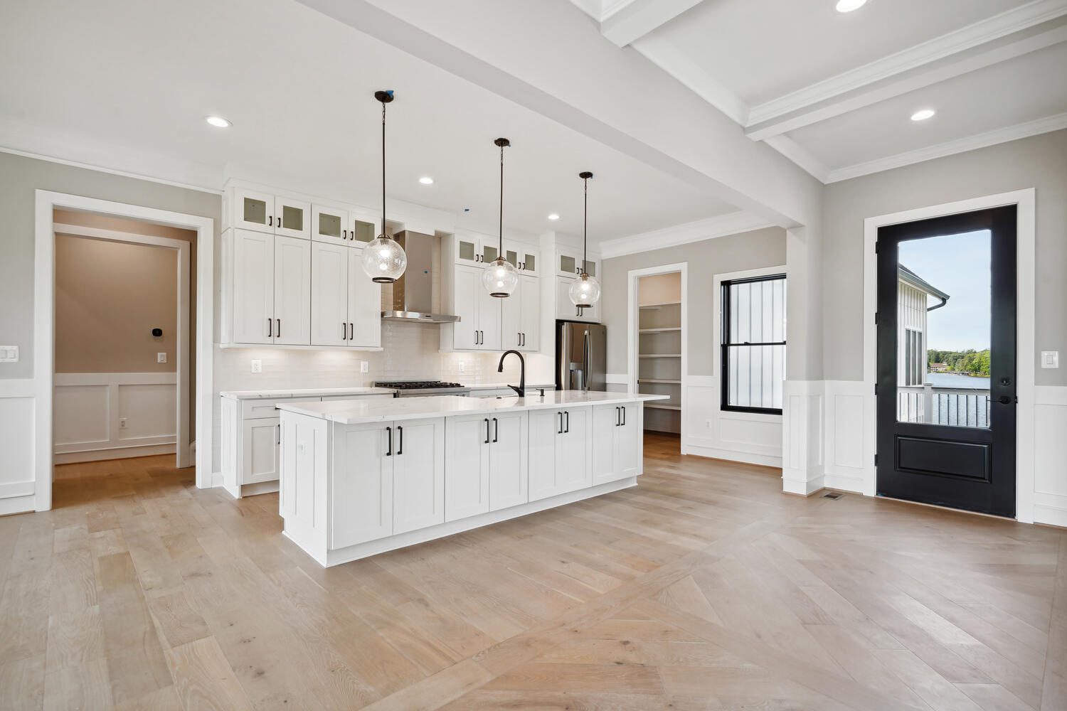 Spacious modern kitchen with white cabinets, an island with pendant lights, stainless steel appliances, hardwood floors, a black door, and large windows.
