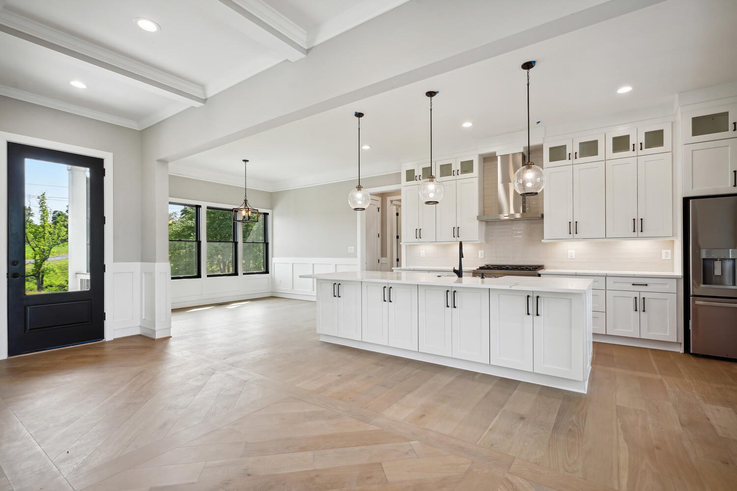Spacious modern kitchen with white cabinets, stainless steel appliances, and pendant lights. Light wood flooring extends to a dining area with large windows and a glass door.
