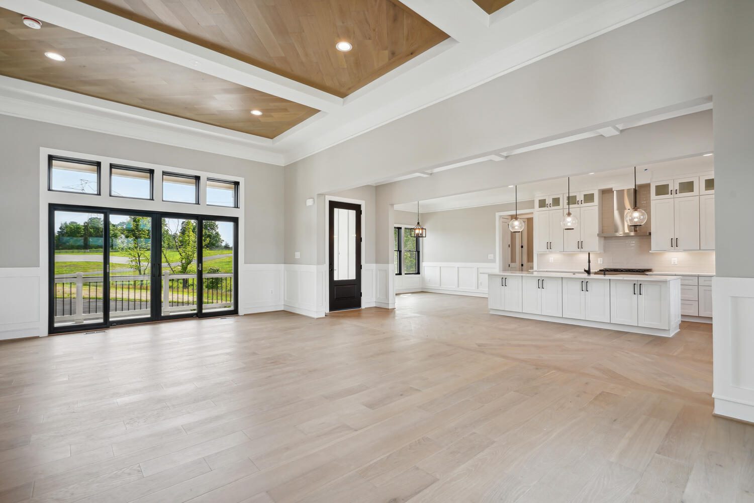 Spacious open-plan living area with light wood floors, a coffered ceiling, large windows, and a modern kitchen with white cabinetry and pendant lights.