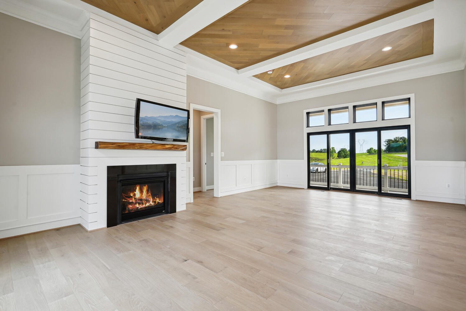 A spacious, empty living room features a modern fireplace, wall-mounted TV, wooden floor, and large windows with a view of greenery.