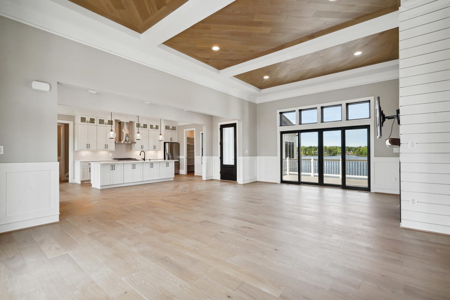 Spacious living area with light wood floors, a modern kitchen, large windows, and a ceiling with wood paneling. A door leads to a balcony with a view.