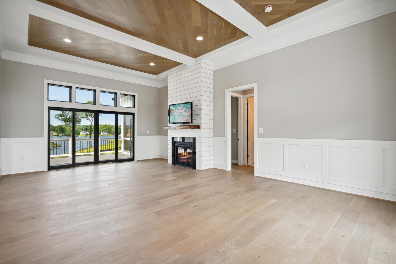 Spacious living room with hardwood floors, fireplace, built-in TV, large windows, and a view of a deck and trees. Ceiling features wood paneling and recessed lighting.