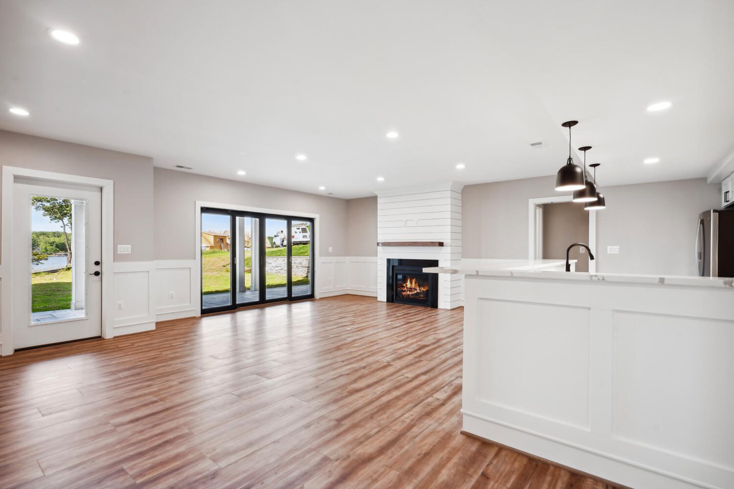 Spacious living area with wooden flooring, a fireplace, large glass doors leading outside, and a kitchen bar with pendant lights.