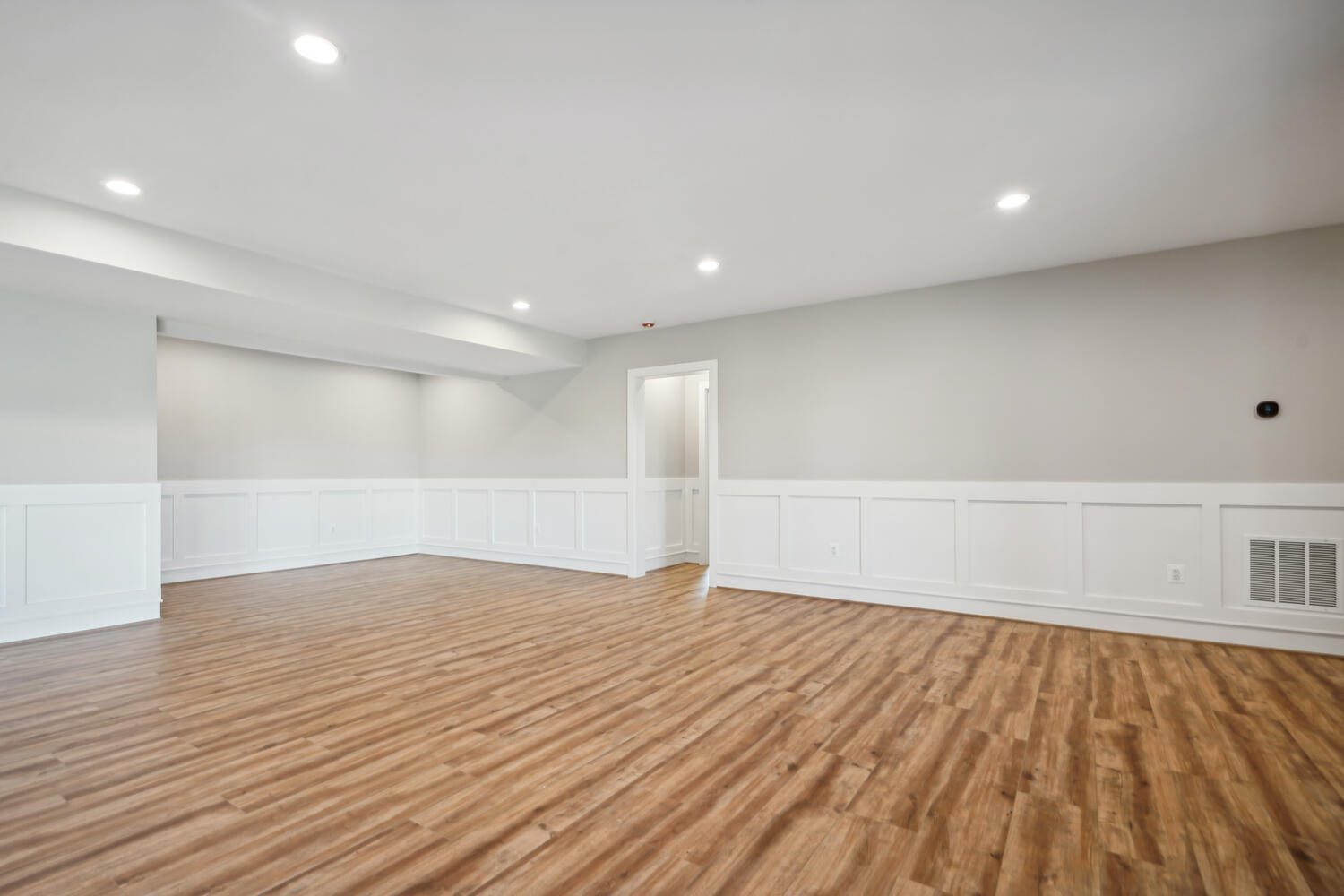 Spacious empty room with light wood flooring, white wainscoting, and neutral-colored walls, lit by recessed ceiling lights.