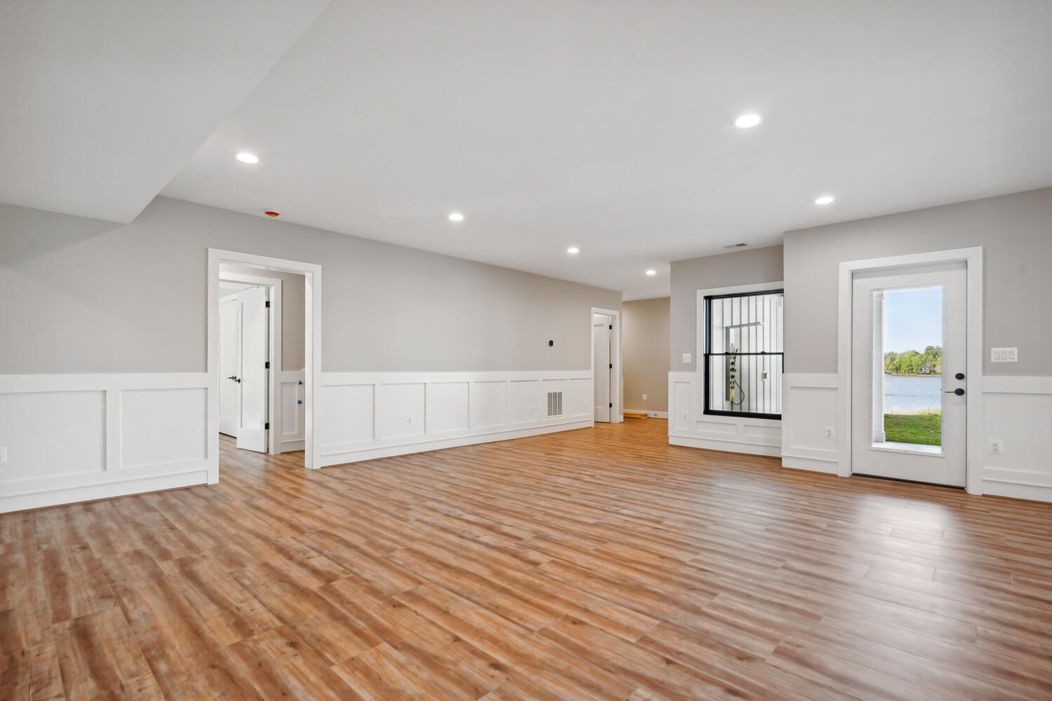 Spacious empty room with wooden floors, white wainscoting, and gray walls. There are two doors and a large window on the right. Recessed ceiling lights illuminate the space.
