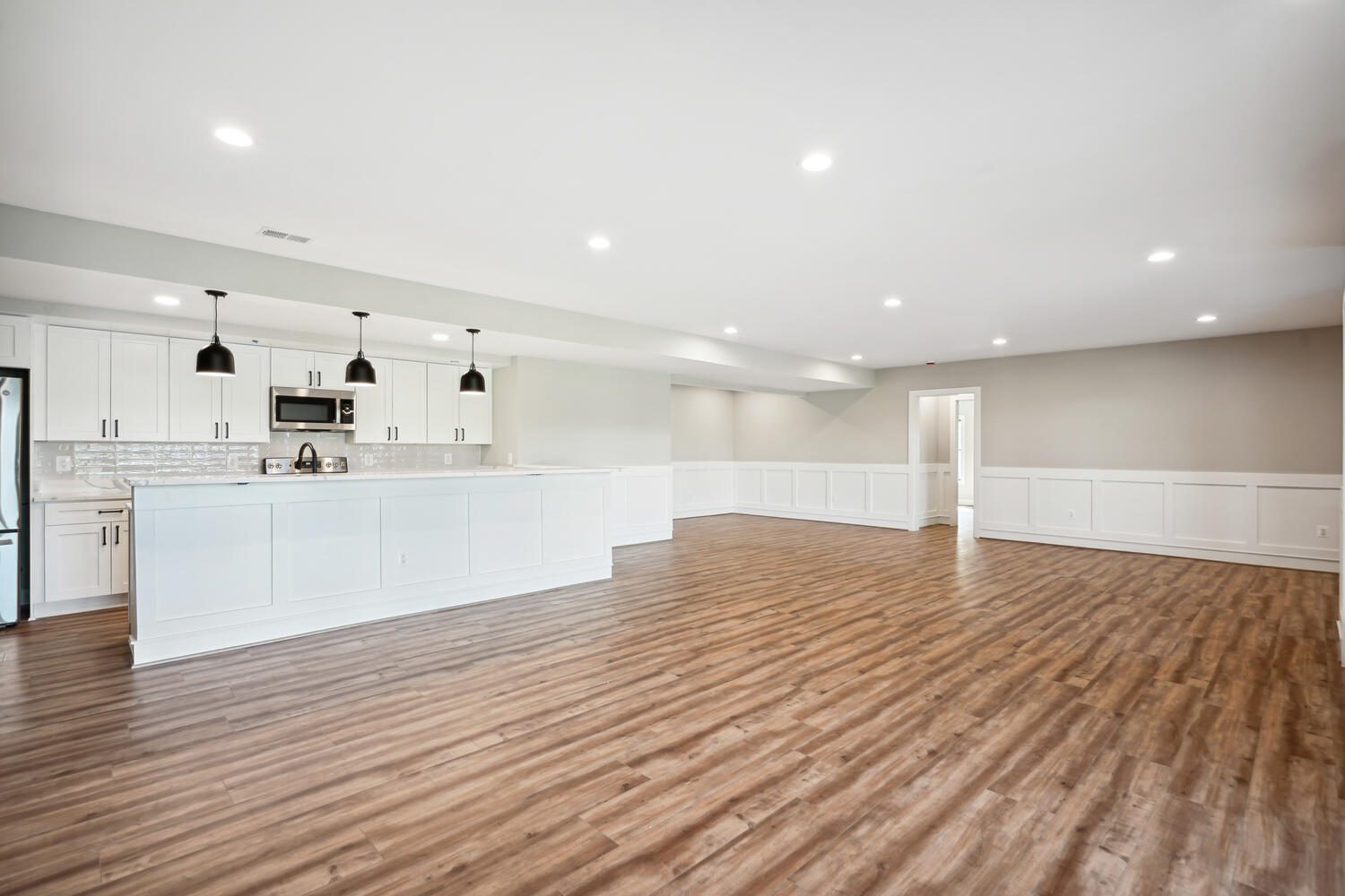 Spacious, empty room with wood flooring and white kitchen cabinets. It features recessed lighting, black pendant lights, and light gray walls.