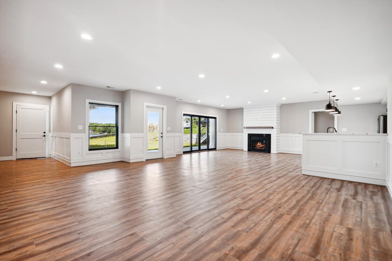 Spacious open-concept living room with wood flooring, recessed lighting, a fireplace, large windows, sliding glass doors, and an adjacent kitchen with a counter and pendant lights.