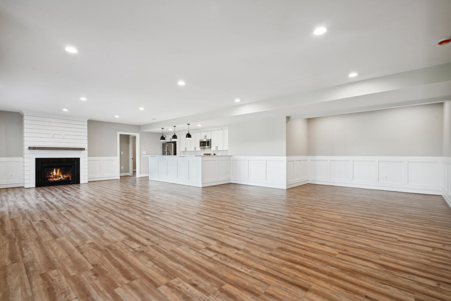 Spacious empty room with wood flooring, recessed lighting, a fireplace, and a view into a modern kitchen. White wainscoting on walls enhances the clean, open layout.