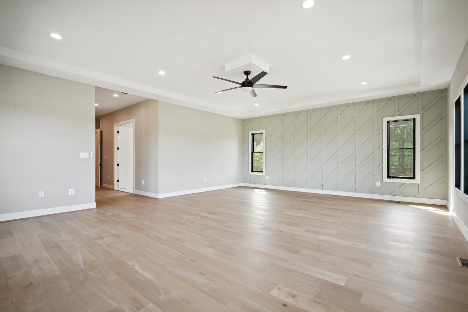 A spacious, empty room with light wood flooring, neutral walls, a patterned accent wall, ceiling fan, and recessed lighting. Two windows are on the accent wall.