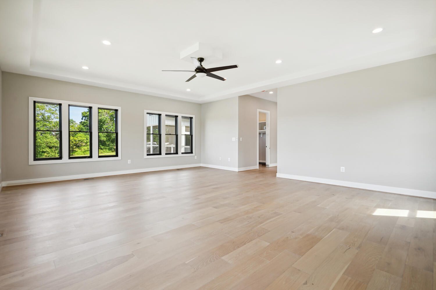 Spacious empty room with large windows, light wood flooring, neutral walls, recessed lighting, and a ceiling fan.