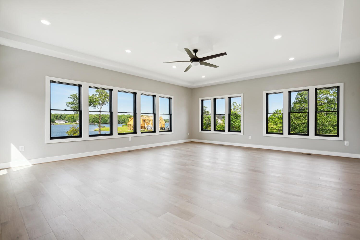 Spacious empty room with light wood flooring, large windows showcasing a lake view, and a ceiling fan.