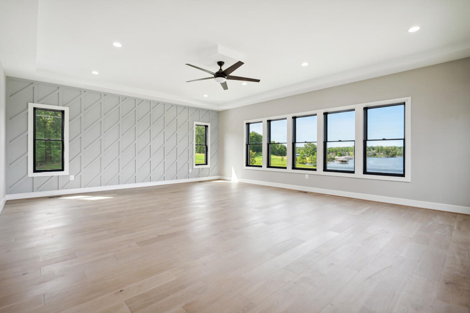 Spacious empty room with light wood flooring, gray accent wall, ceiling fan, and large windows overlooking a scenic outdoor view.