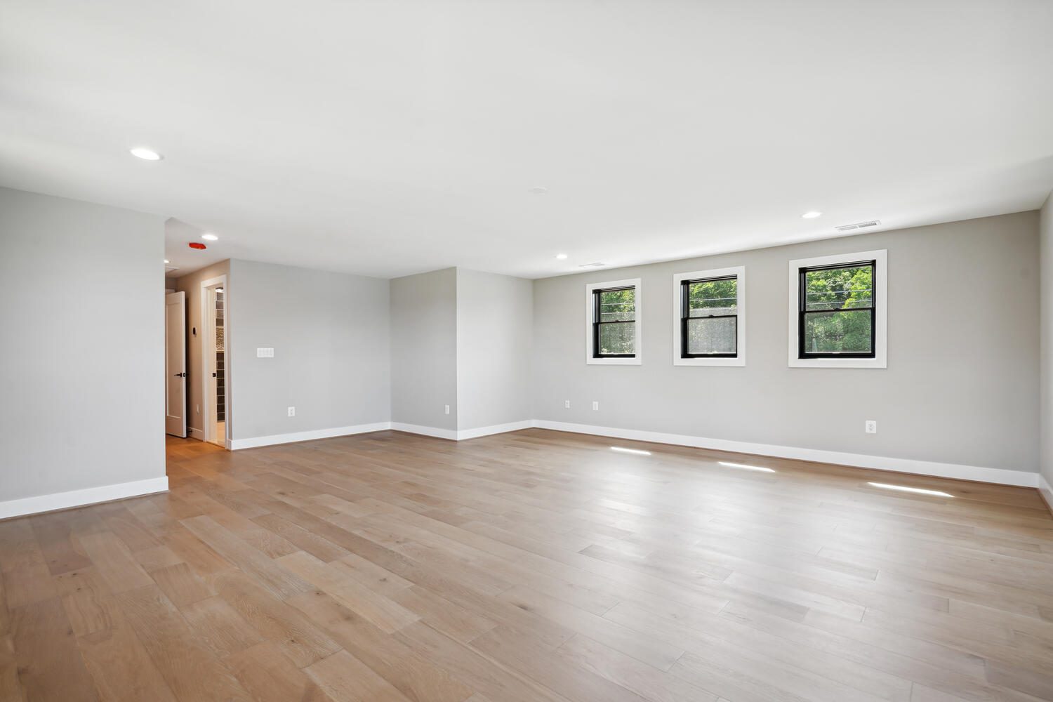 Empty room with light wood flooring, three windows, and gray walls. A doorway on the left leads to an adjacent area. Ceiling lights are evenly spaced.