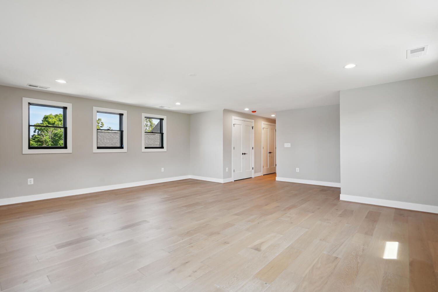 Spacious empty room with light wood flooring, three small windows, white walls, and recessed lighting. A hallway with closed doors is visible on one side.