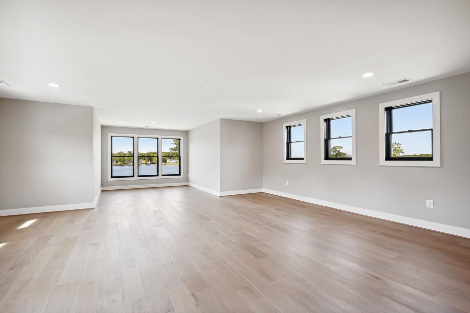 Spacious empty room with light wooden flooring, three small windows on the right, and large glass doors leading to a balcony. Walls are painted light gray.