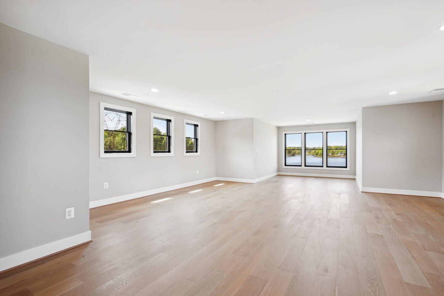 Spacious, empty room with wooden floors, neutral walls, and large windows offering a view of greenery and water outside.