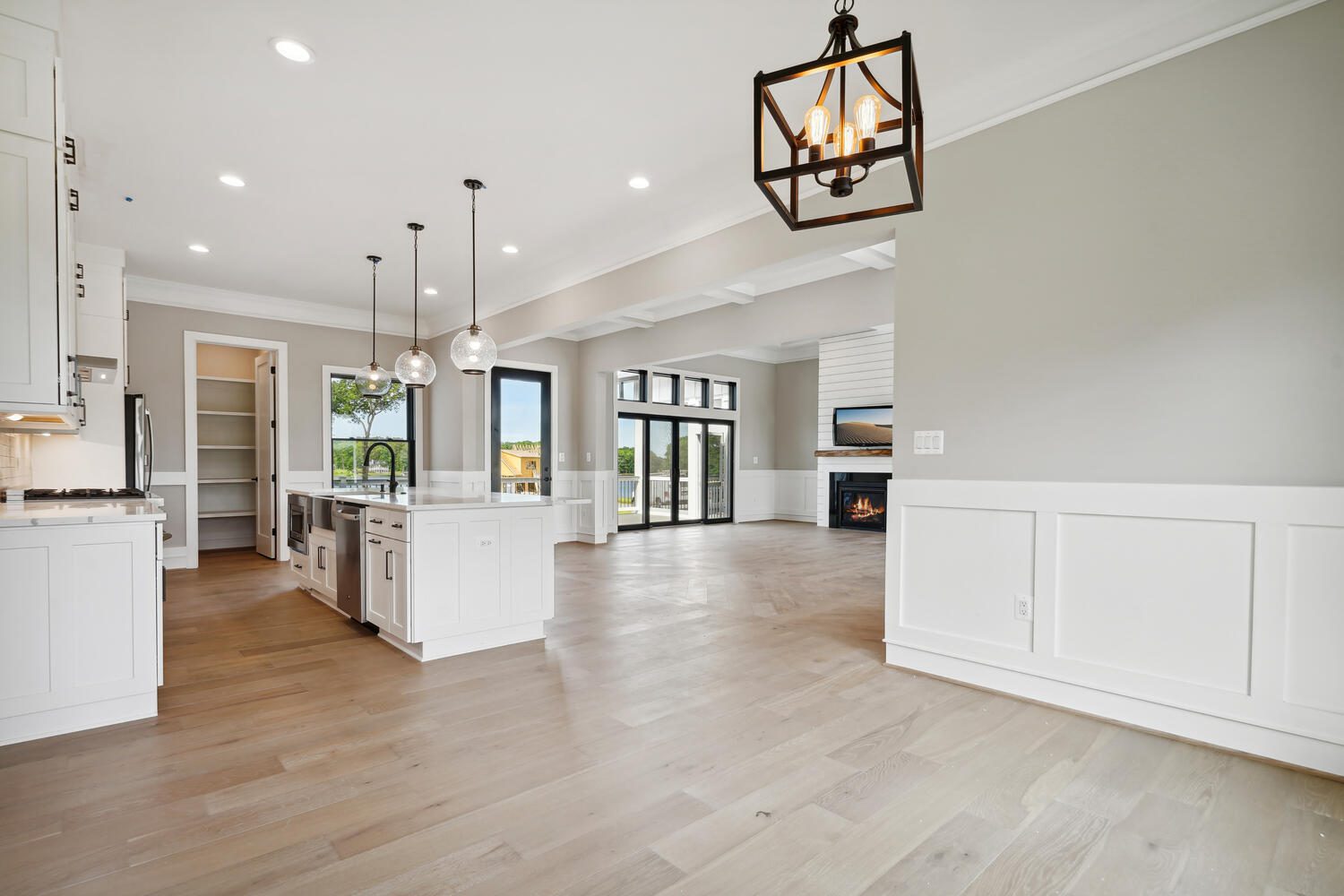 Spacious, modern kitchen and living area with white cabinetry, wooden flooring, large windows, and pendant lighting. A fireplace is visible in the living area.