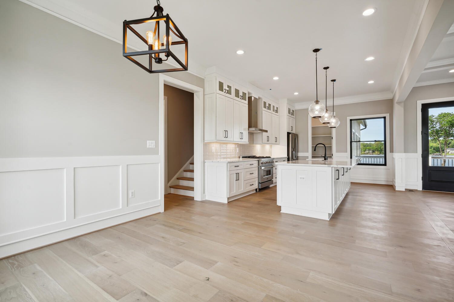 Spacious kitchen with white cabinetry, large island, modern pendant lights, wood flooring, and stainless steel appliances. Bright natural light from windows.