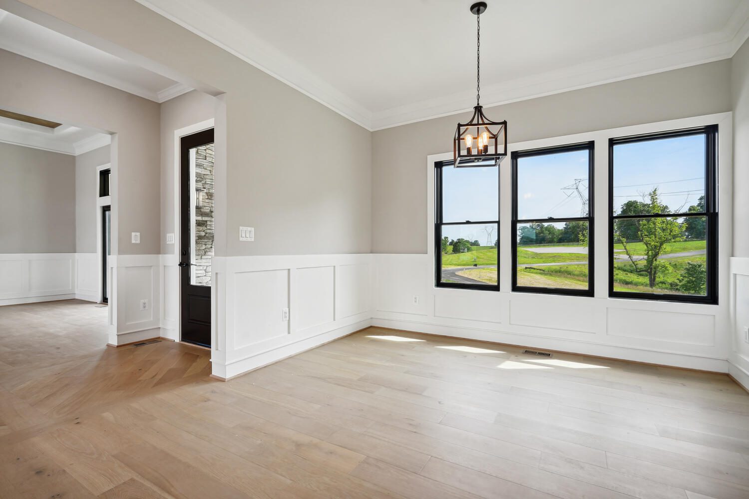 An empty room with light wood flooring, white wainscoting, and large windows. A black pendant light hangs from the ceiling, and a partially open door leads to another room.