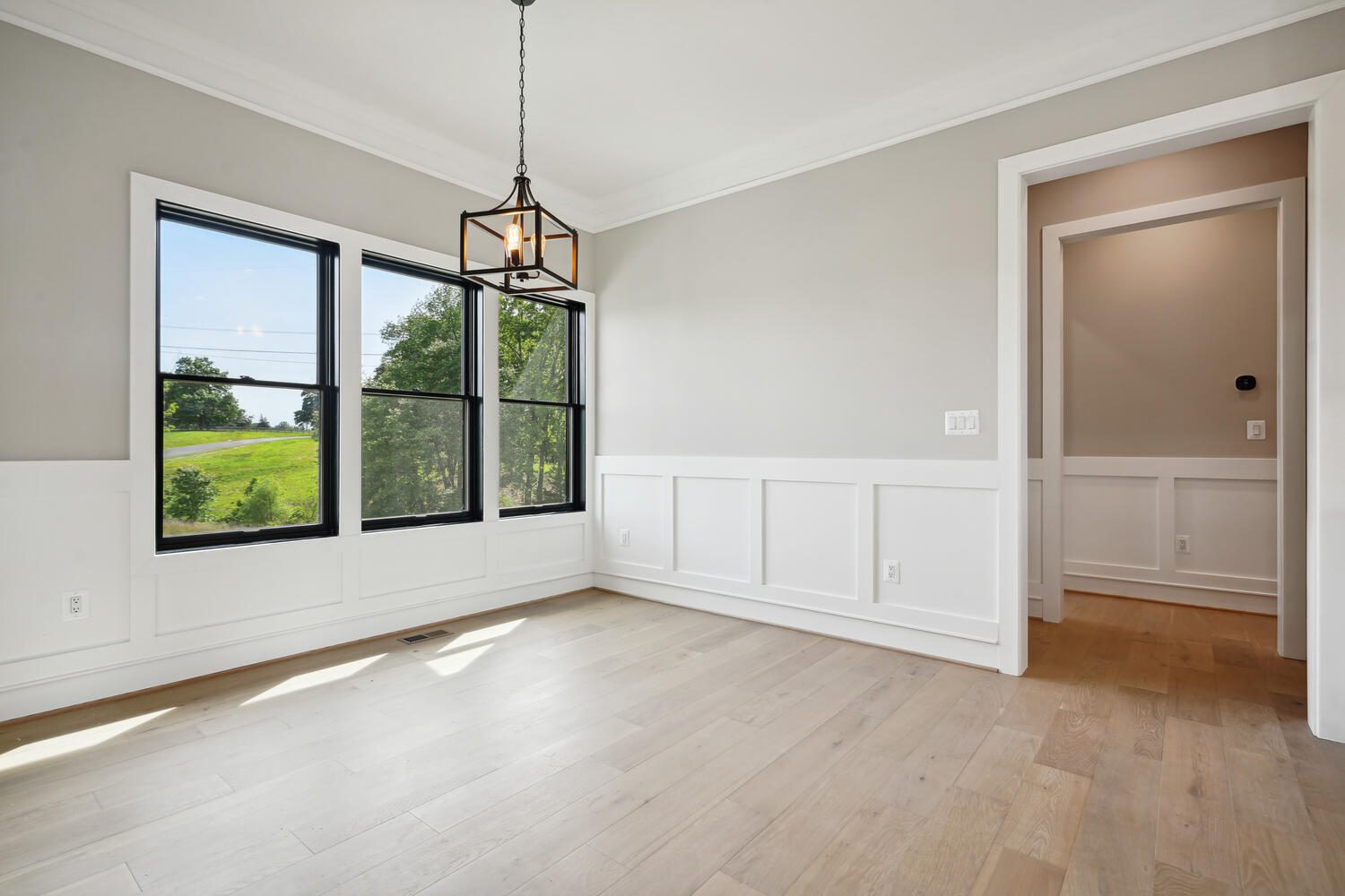 Bright, empty room with light wood flooring, white wainscoting, and large windows showing a green landscape. A black-framed light fixture hangs from the ceiling.