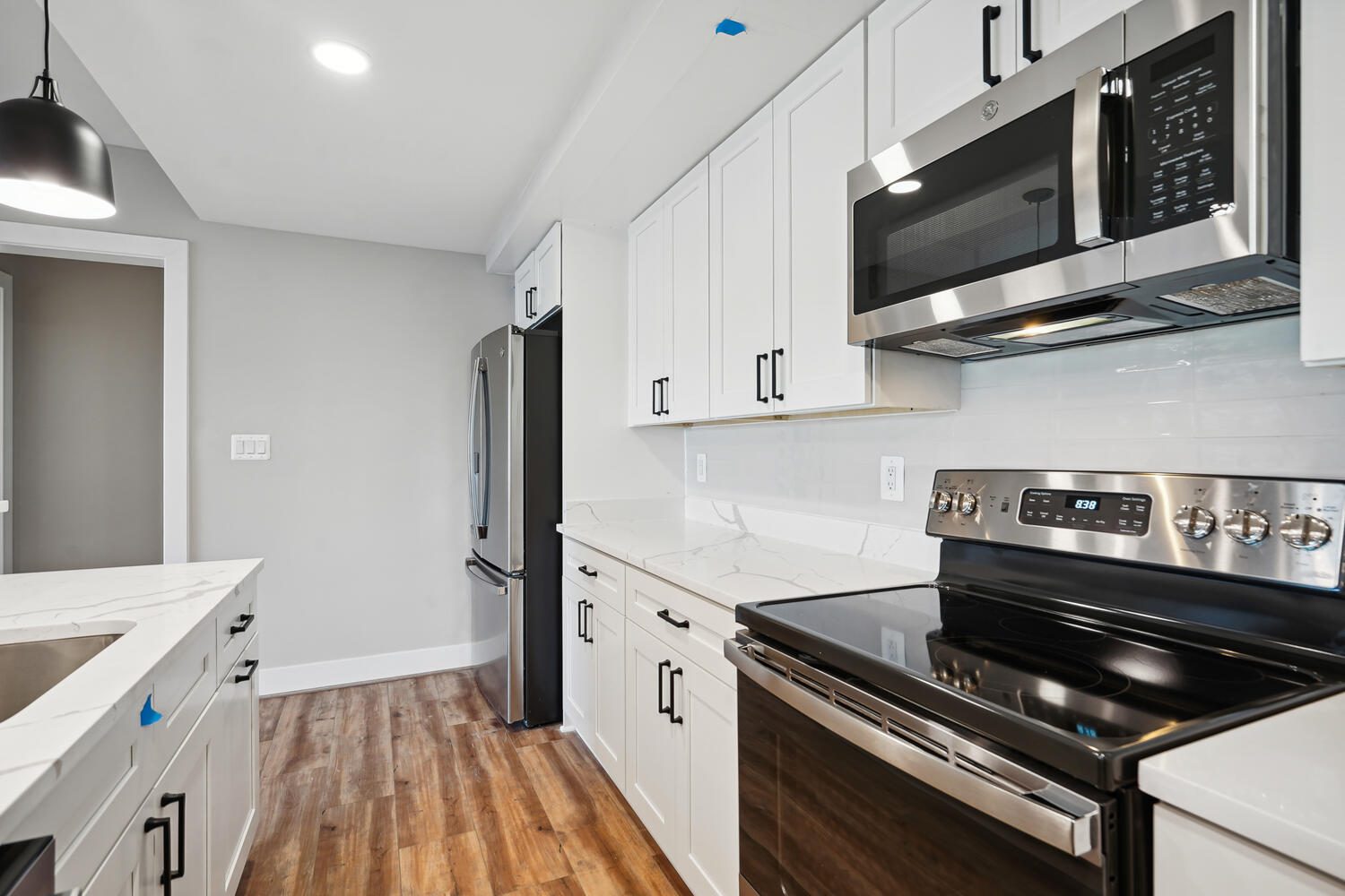 Modern kitchen with stainless steel appliances, hardwood floor, white cabinets, and gray walls. Features an oven, microwave, refrigerator, and a black pendant light over the countertop.