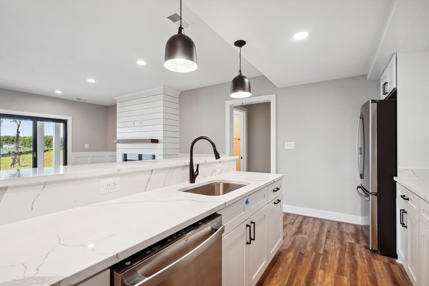 Modern kitchen with a white marble island, stainless steel appliances, black pendant lights, and wood flooring. There's a view of a bright room with large windows in the background.