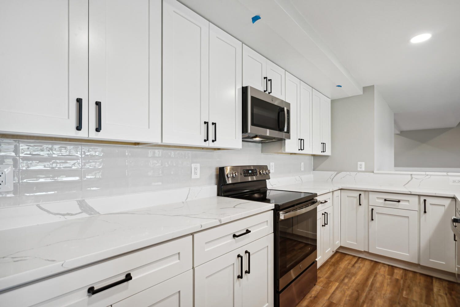Modern kitchen with white cabinets, marble countertops, stainless steel oven, and microwave. Wood flooring and white tiled backsplash are featured.