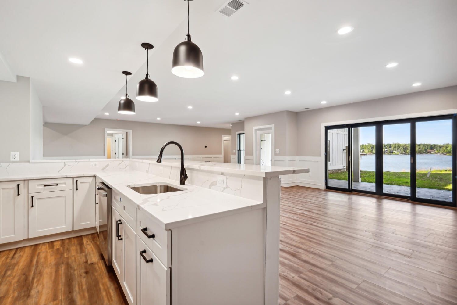 Modern open-plan kitchen with white cabinets, marble countertops, and stainless steel fixtures. Pendant lights hang above island. Large glass doors offer a view of a lake and wooded area.