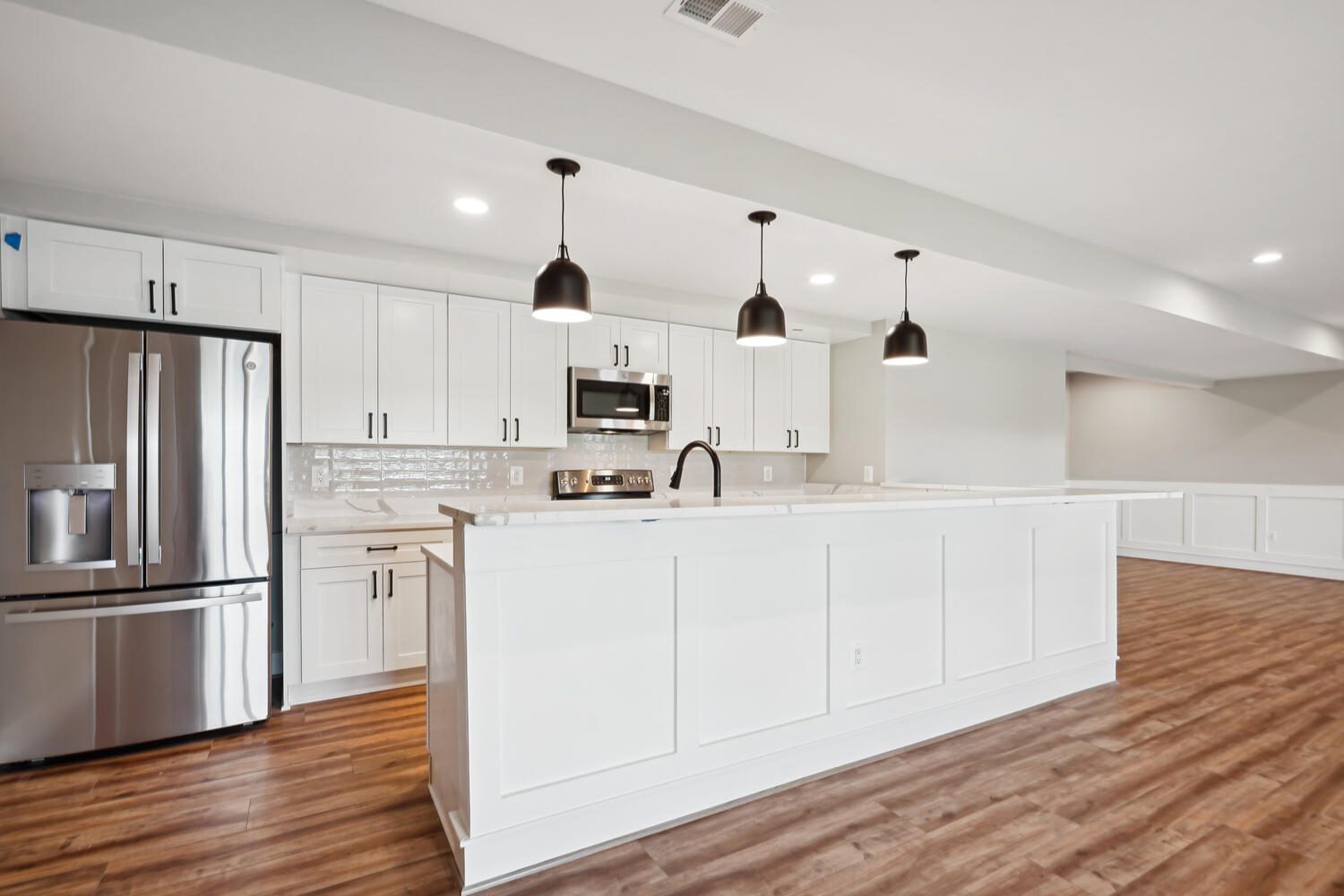 Modern kitchen with white cabinets, a spacious island with a sink, stainless steel appliances, and three black pendant lights above. The floor is wooden and the ceiling has recessed lighting.
