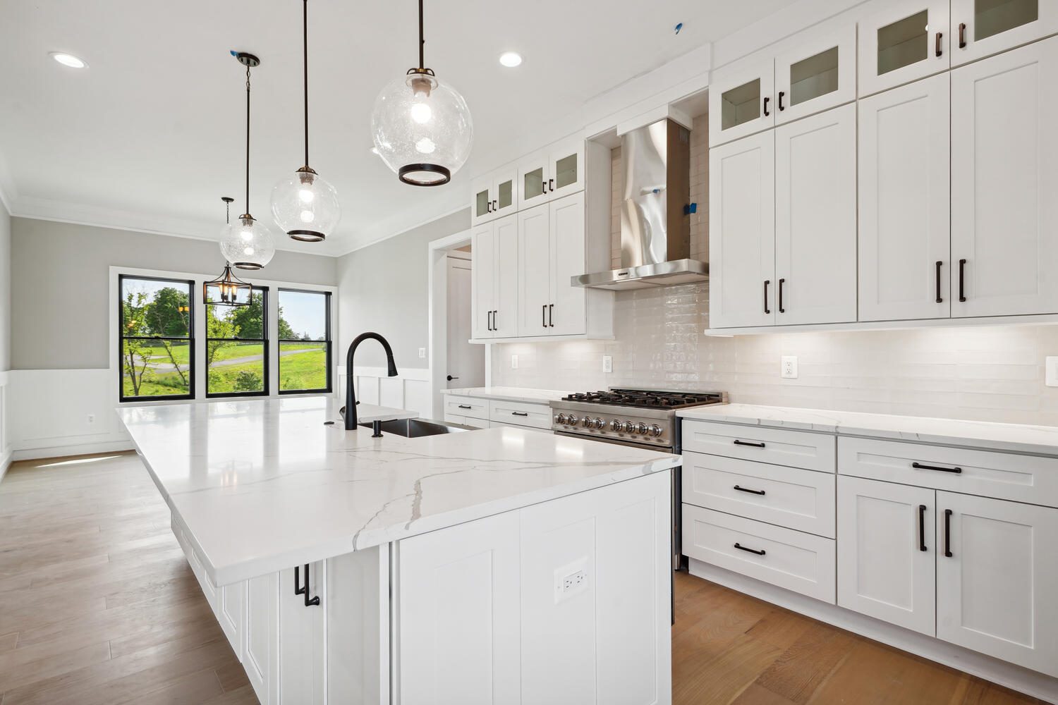 Modern kitchen with white cabinets, marble countertops, pendant lights, stainless steel appliances, and a view of greenery through large windows.