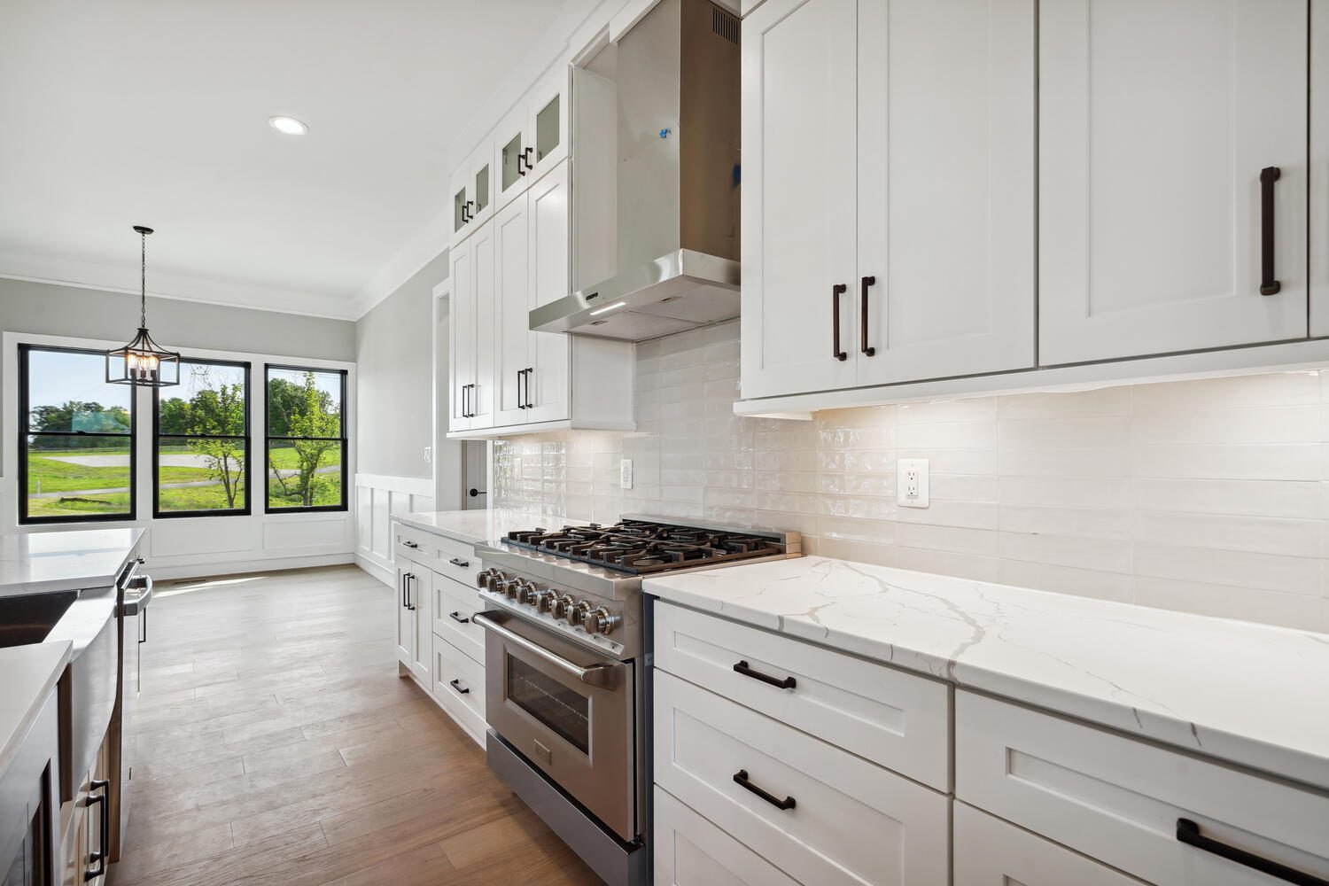 Modern kitchen with white cabinets, stainless steel stove, and marble countertops. Large window offers natural light. Wood flooring complements the clean design.