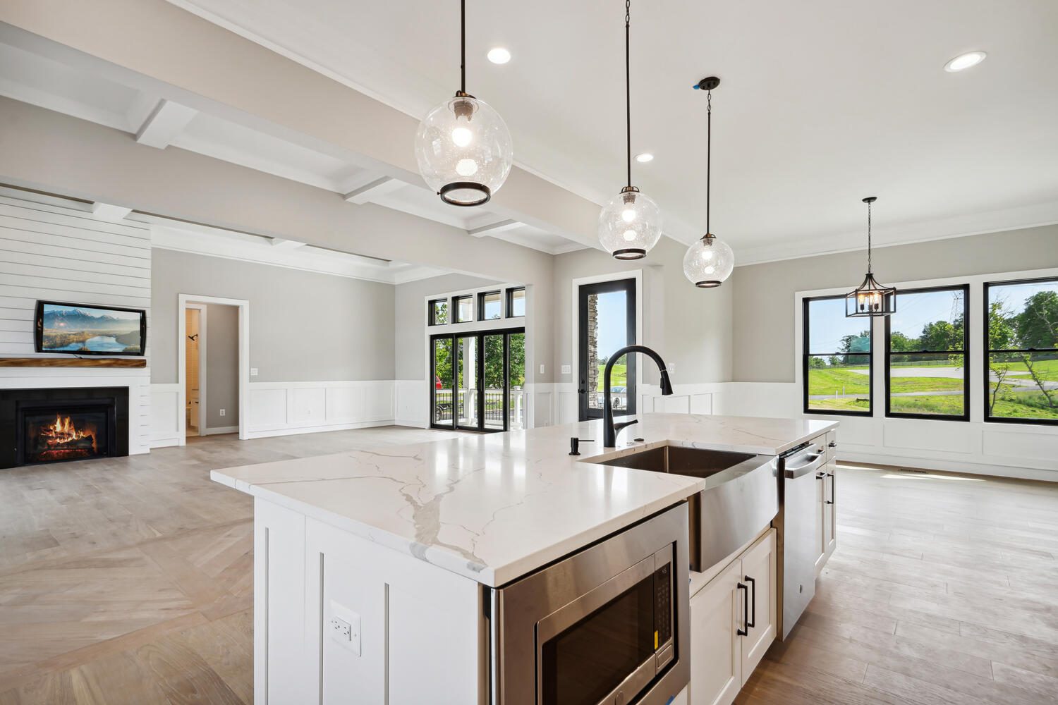 Spacious modern kitchen with marble island, pendant lights, and large windows. Living area has a fireplace, wooden floors, and a view of the outdoors.