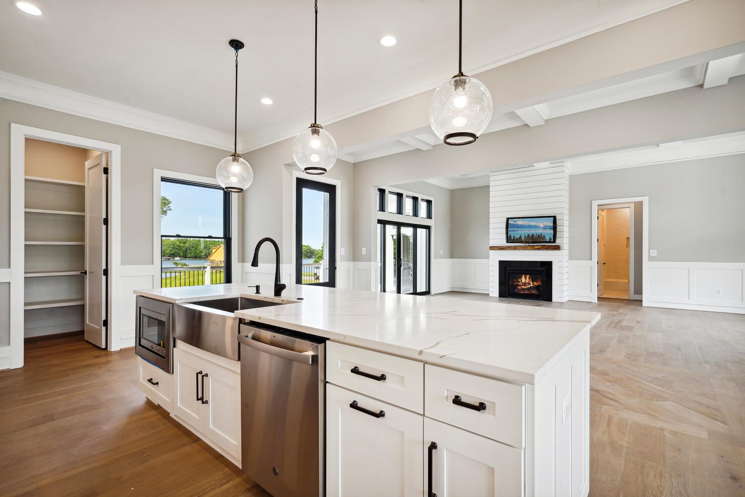 Modern kitchen and living area with white cabinetry, marble countertops, stainless steel appliances, pendant lights, a fireplace, and large windows overlooking greenery.