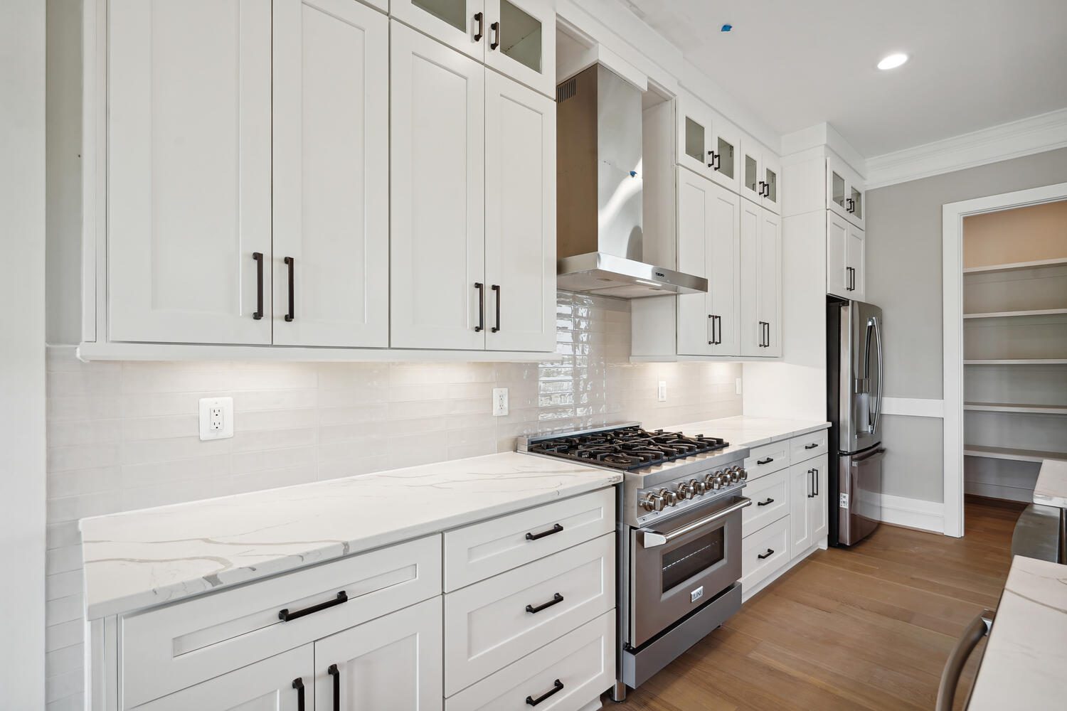 Modern kitchen with white cabinets, marble countertops, a stainless steel stove with a vent hood, and hardwood floors. A walk-in pantry is visible on the right.