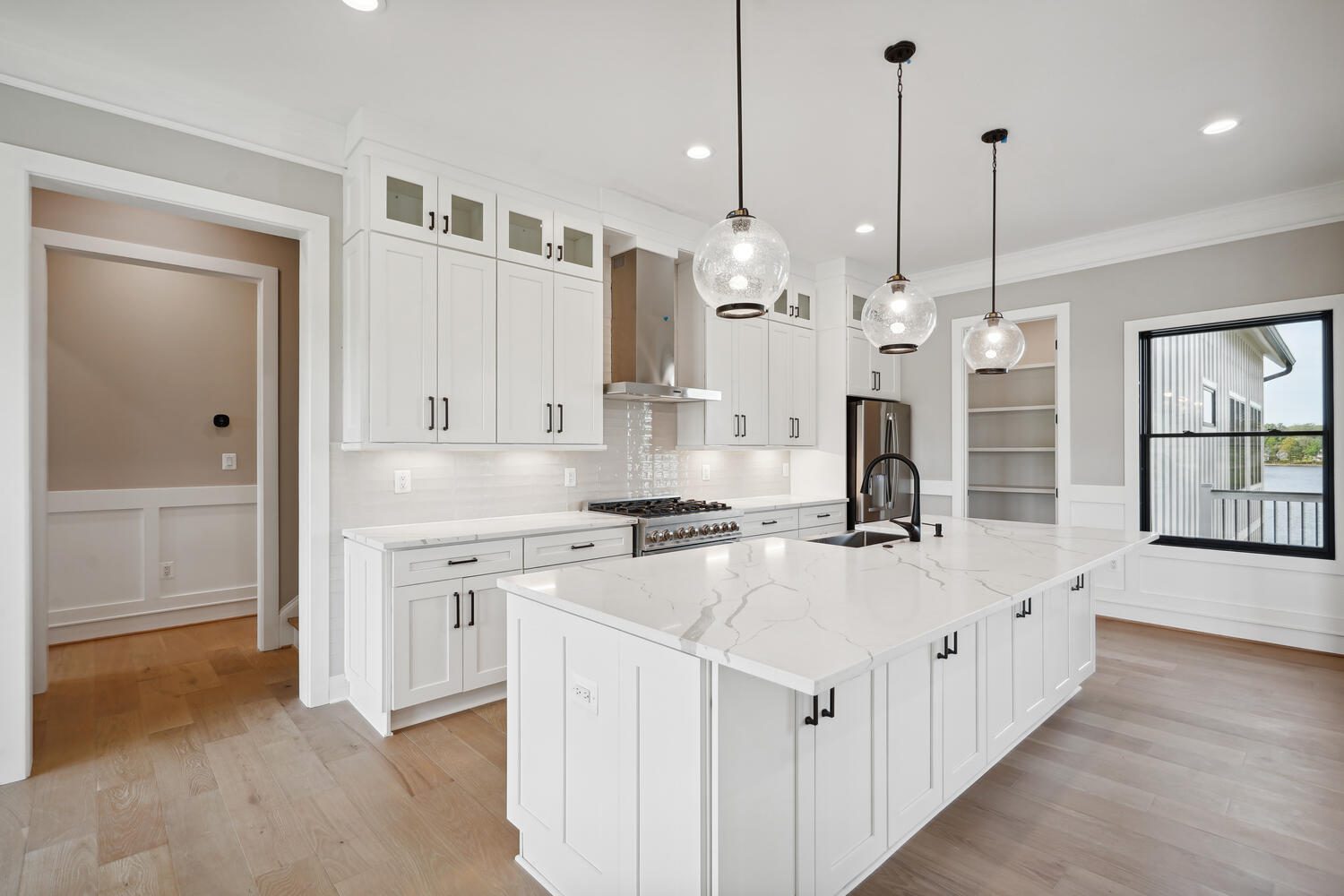 Modern kitchen with white cabinetry, marble island, pendant lights, and stainless steel appliances. Light wood flooring complements the bright, open space.