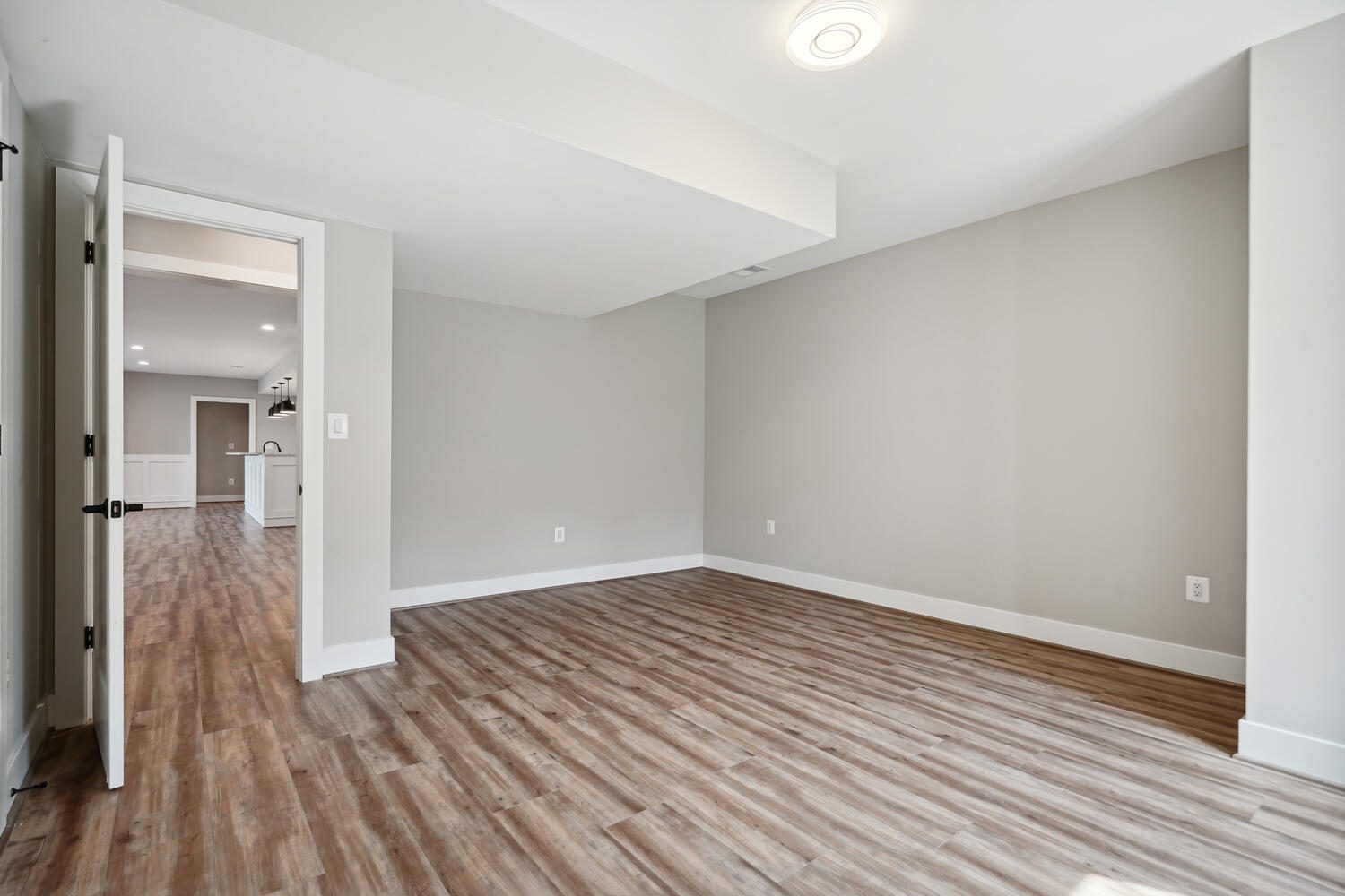 Empty room with light gray walls, wood flooring, and a white ceiling. An open door reveals a view of a kitchen area in the background.