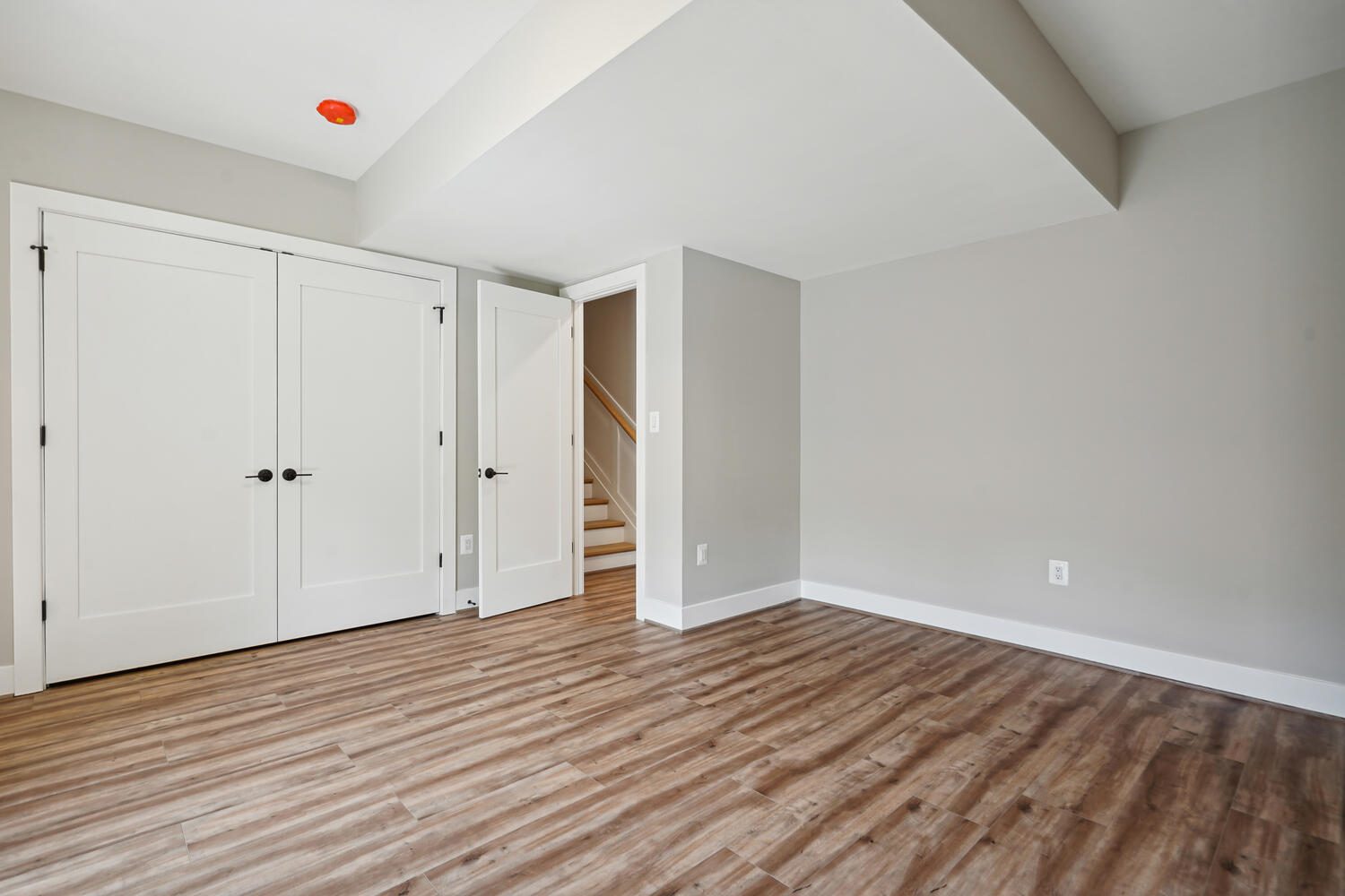 Empty room with light gray walls and wood flooring. White double doors and a white single door lead to a stairway. A small ceiling-mounted smoke detector is visible.